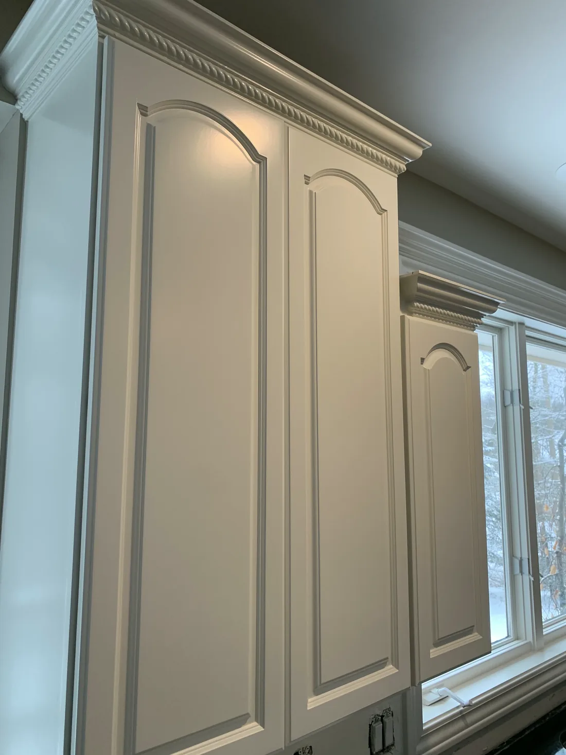 Close on a run of arched cabinet doors and crown moulding, freshly finished, beside a window.