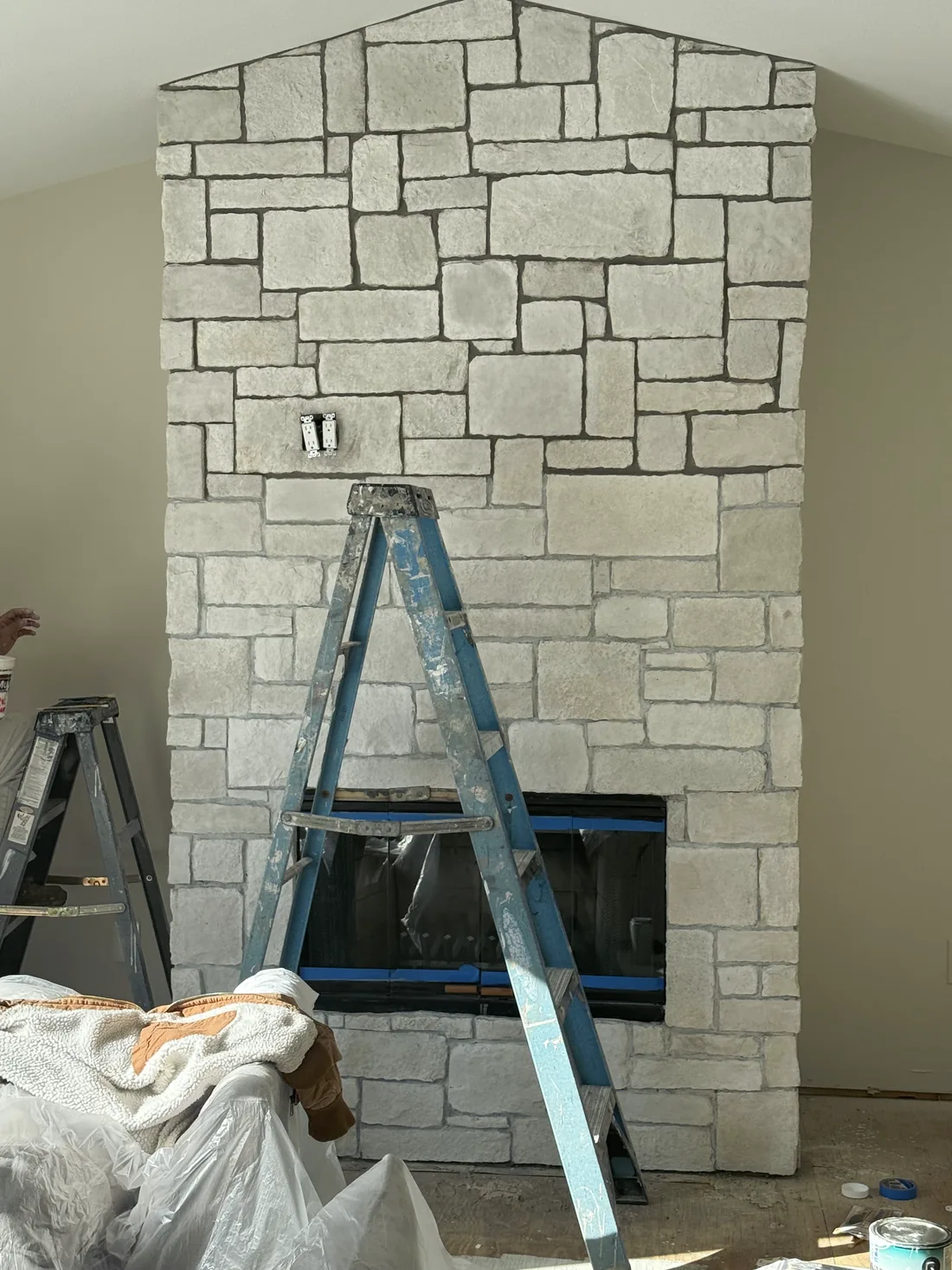A stone fireplace mid-job with a ladder in front of it and drop sheets on the floor.
