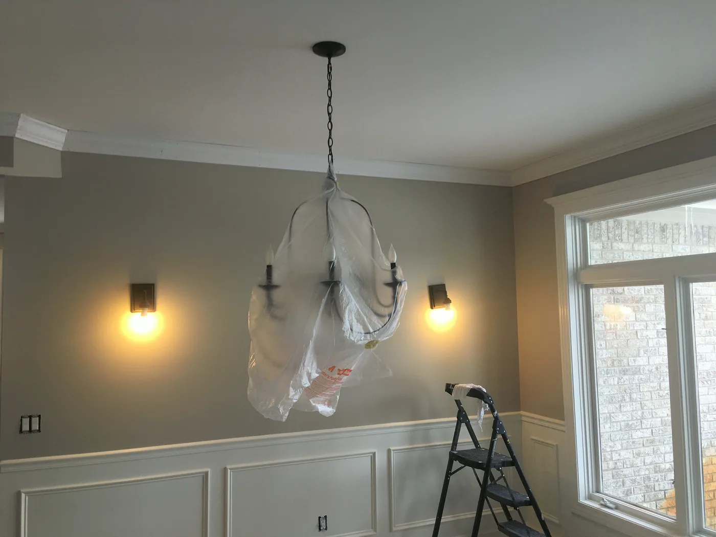 A dining room mid-job: the chandelier wrapped in plastic, wainscoting taped, a step ladder by the window.